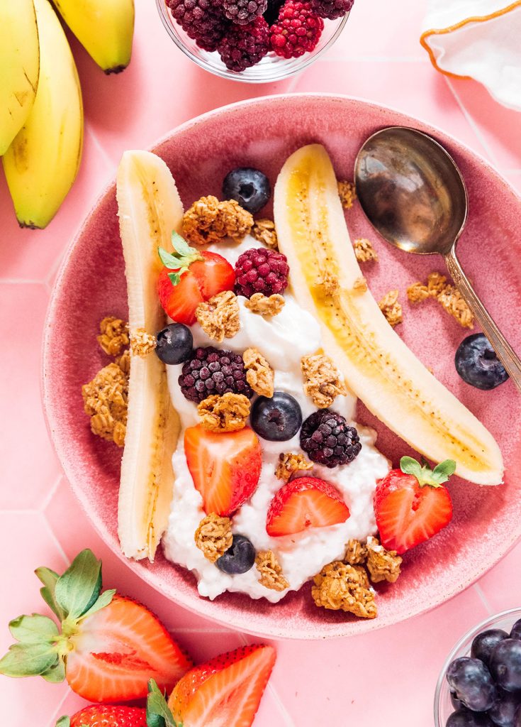 A banana split in a pink bowl with berries, granola, and yogurt with a spoon