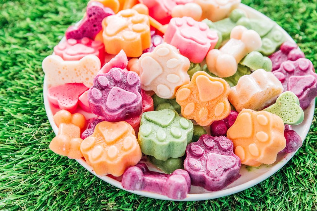 A bowl of multi colored frozen dog treats in the shape of paws and bones.