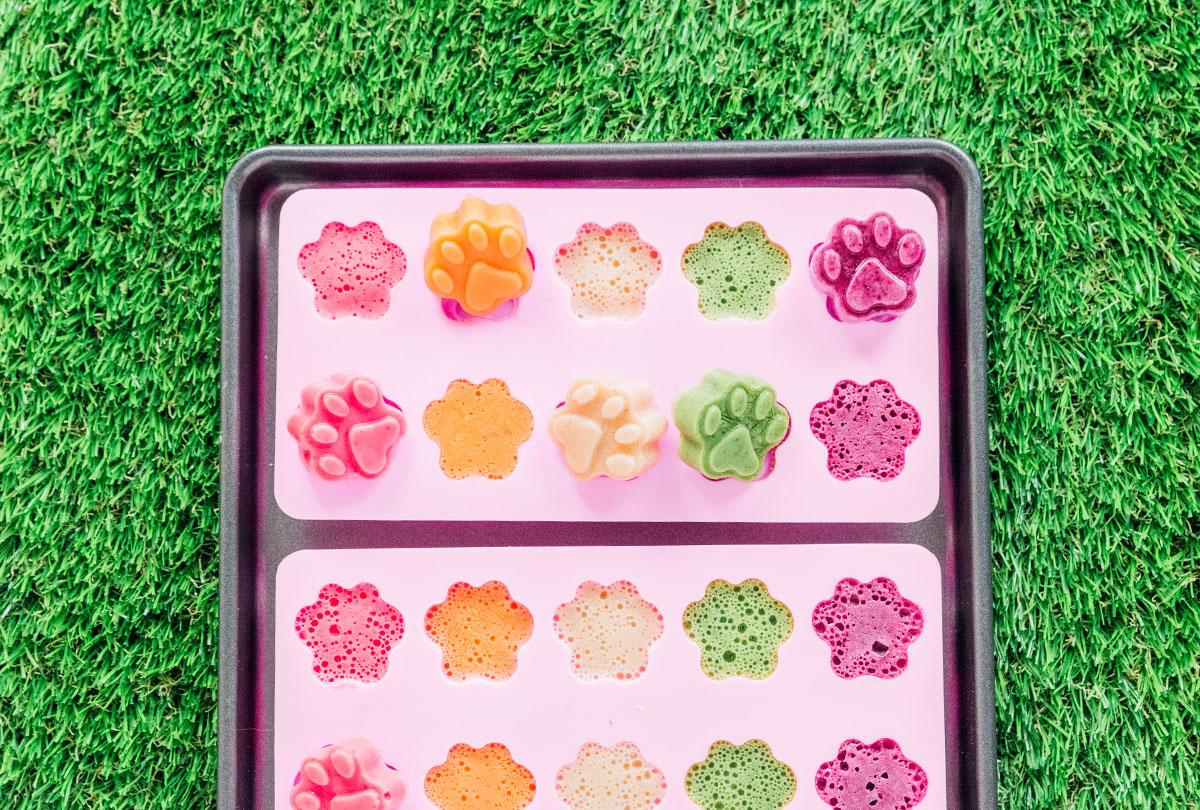 A sheet pan with two paw print molds laid flat and filed with strawberry, carrot, banana, kale, and blueberry frozen treat mixture.